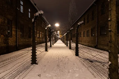 Fenced lane between blocks on a snowy night, Auschwitz I, Poland — Nicolas Marincic